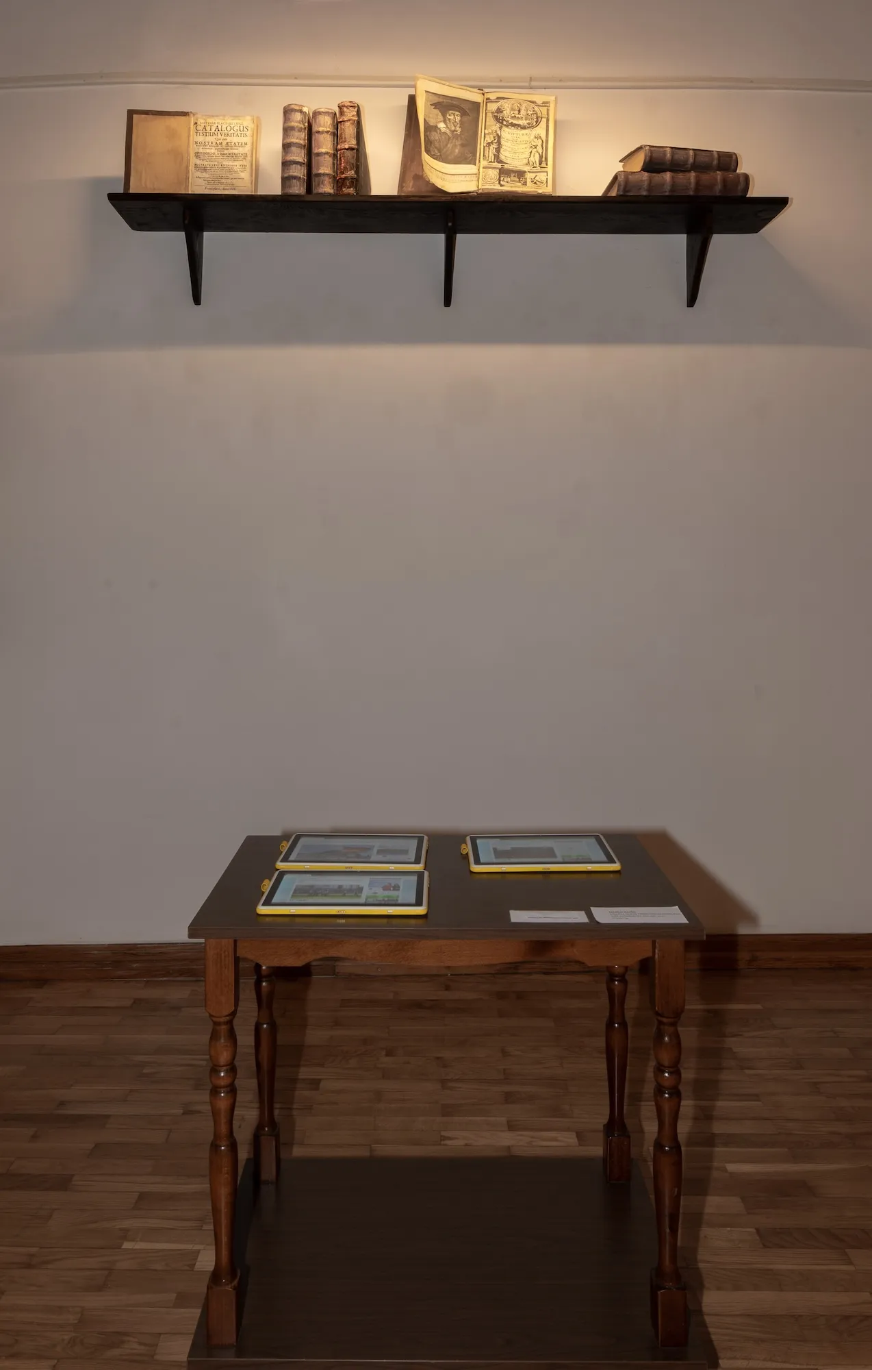 Installation view of a wall shelf holding antique books above a table with three tablets displaying news articles.