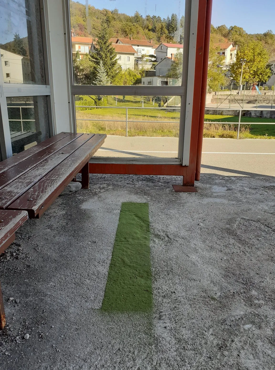 A strip of green linden-leaf powder on the concrete floor of a bus shelter, with a wooden bench and a village view through the glass behind it.