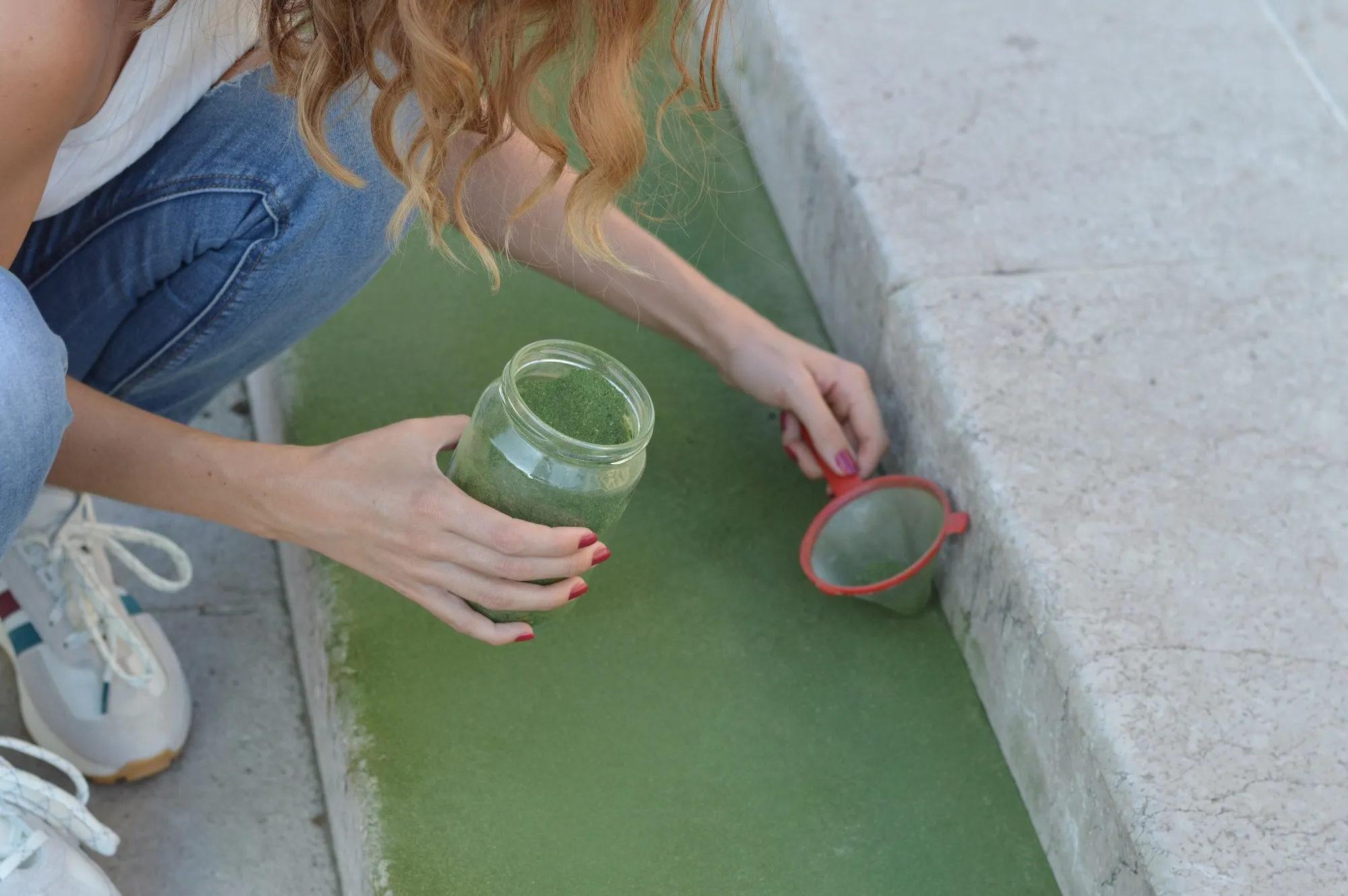 The artist kneeling beside a fountain, straining green linden-leaf powder through a small sieve onto the pavement.