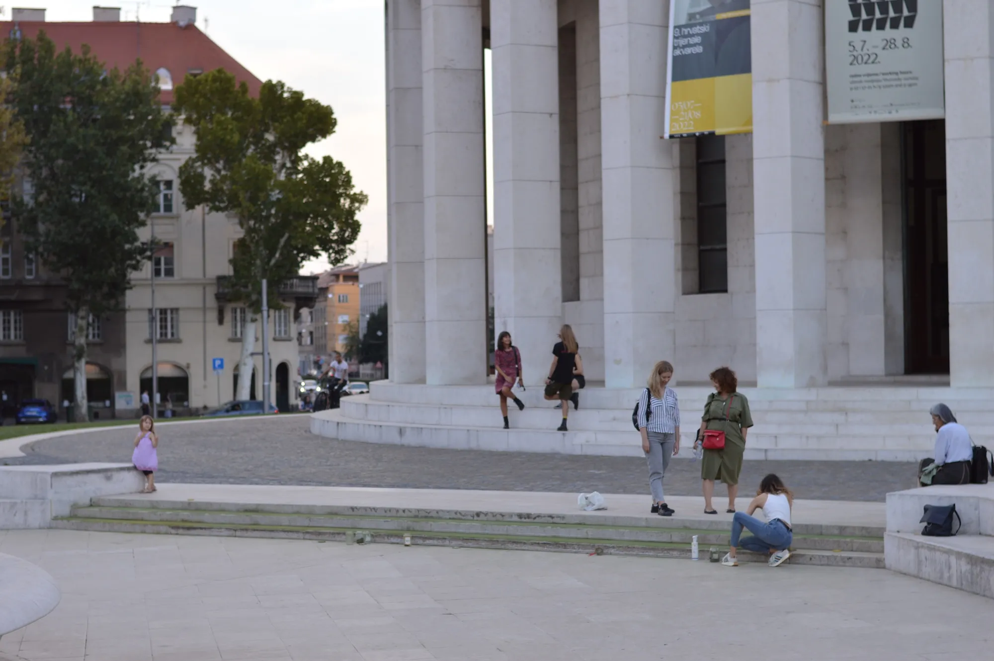 A man crouching to pet two small dogs near the fountain, with a child running past.