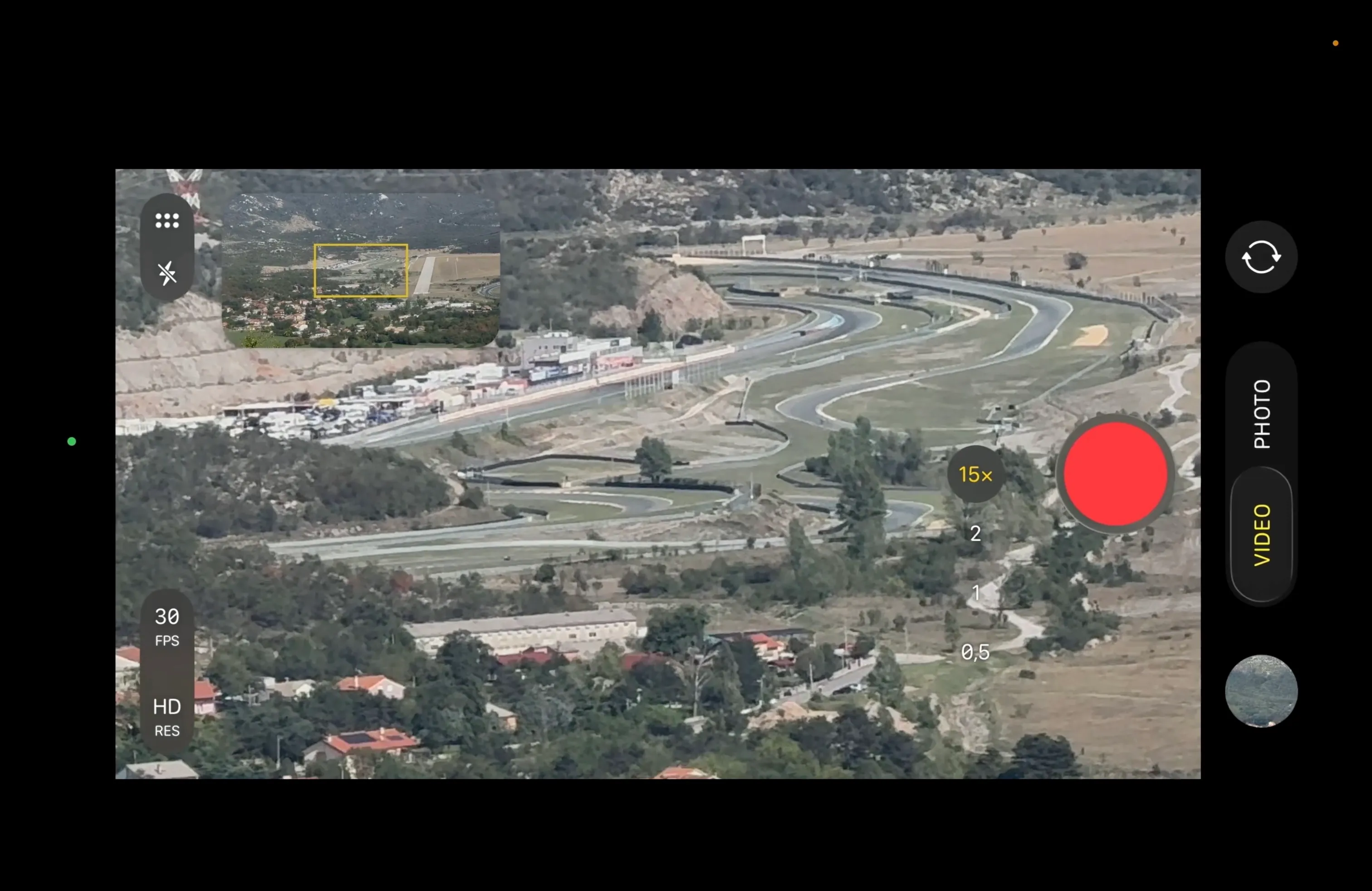 A screen-recorded phone camera interface at 15× zoom, showing the winding track of Grobnik Circuit with parked vehicles along the pit lane and houses in the foreground.