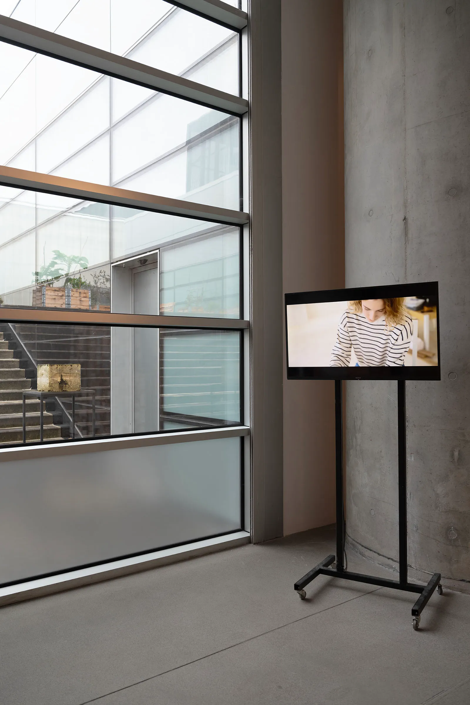 Gallery installation view with a video screen on a stand beside a floor-to-ceiling window, the sculpture visible through the Museum window. Sculpture is displayed outside the gallery 2 meters from the Museum window and video screen inside.