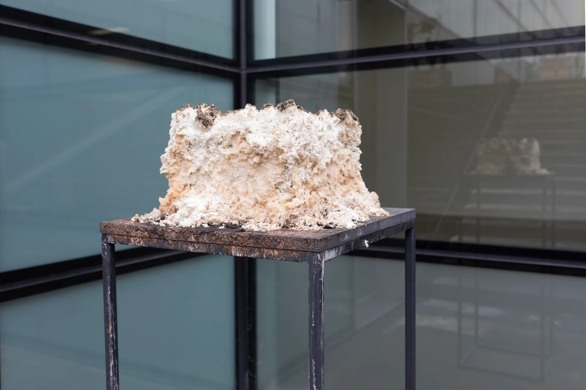 Wide view of the sculpture on its stand in a glass-walled corner, with another artwork faintly visible in the window's reflection.