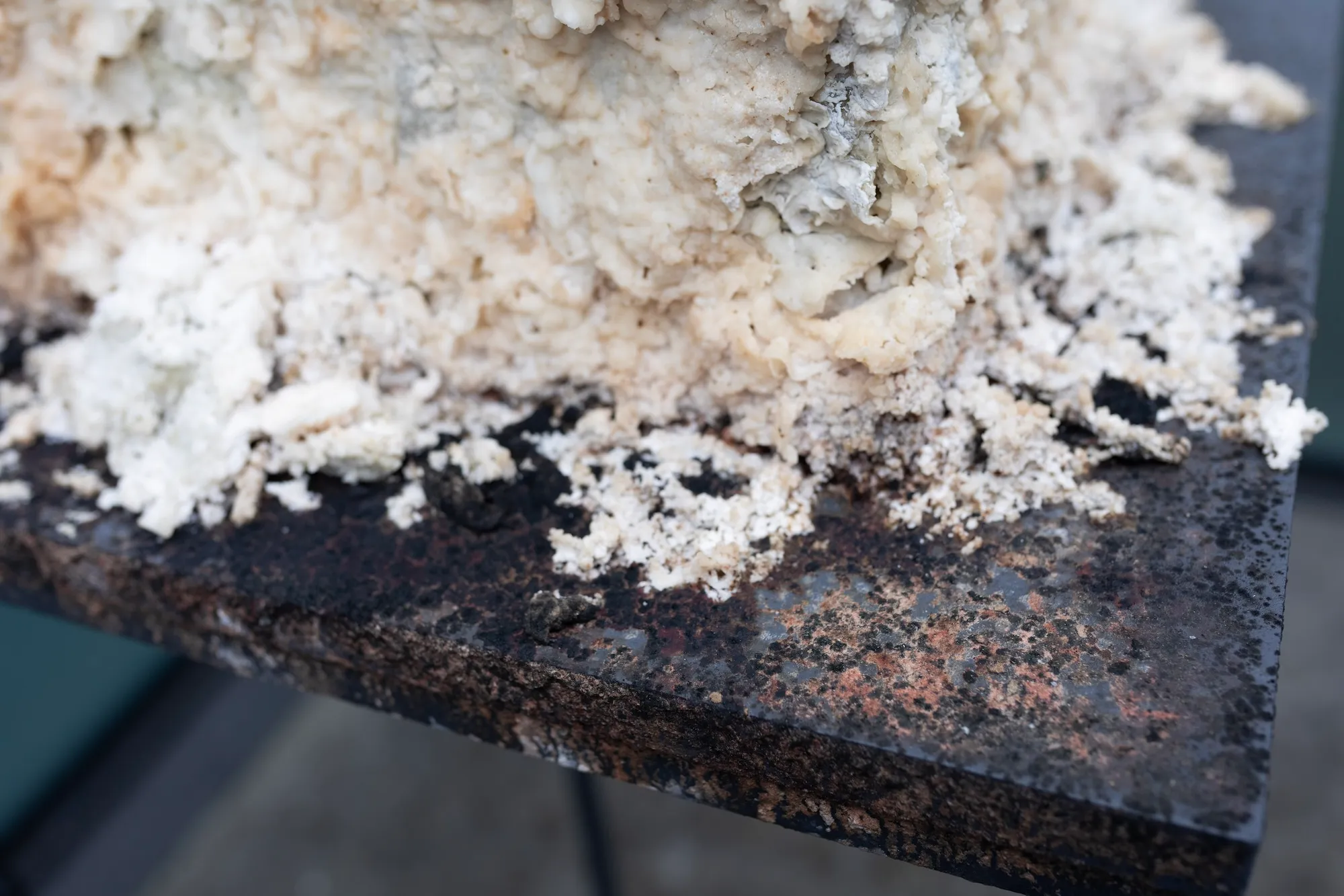 Extreme close-up of the sculpture's pale granular surface meeting the corner of its rusted metal stand.