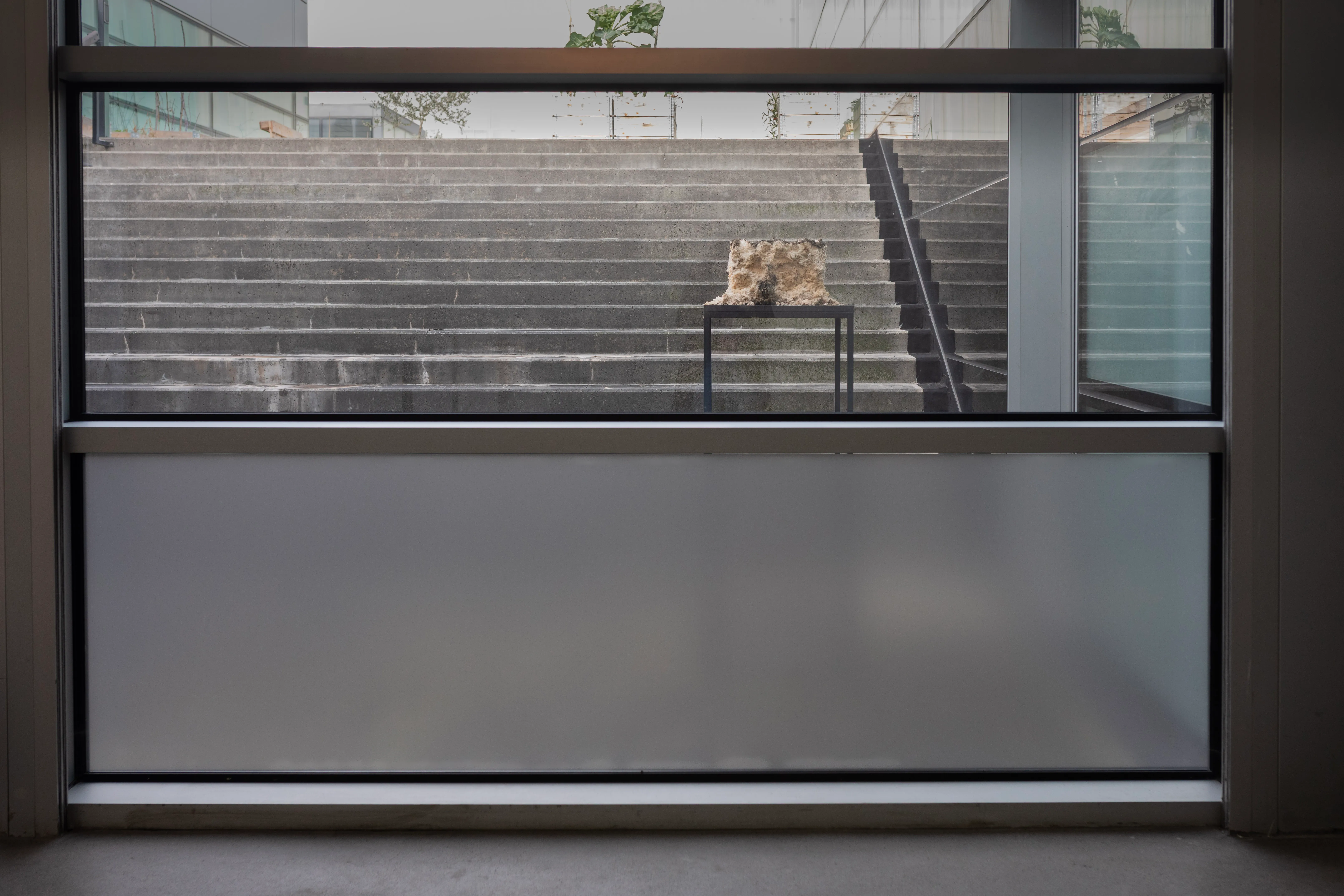 Wide view from inside the building, looking out through a glass wall at the sculpture displayed on its stand against the concrete stairs.