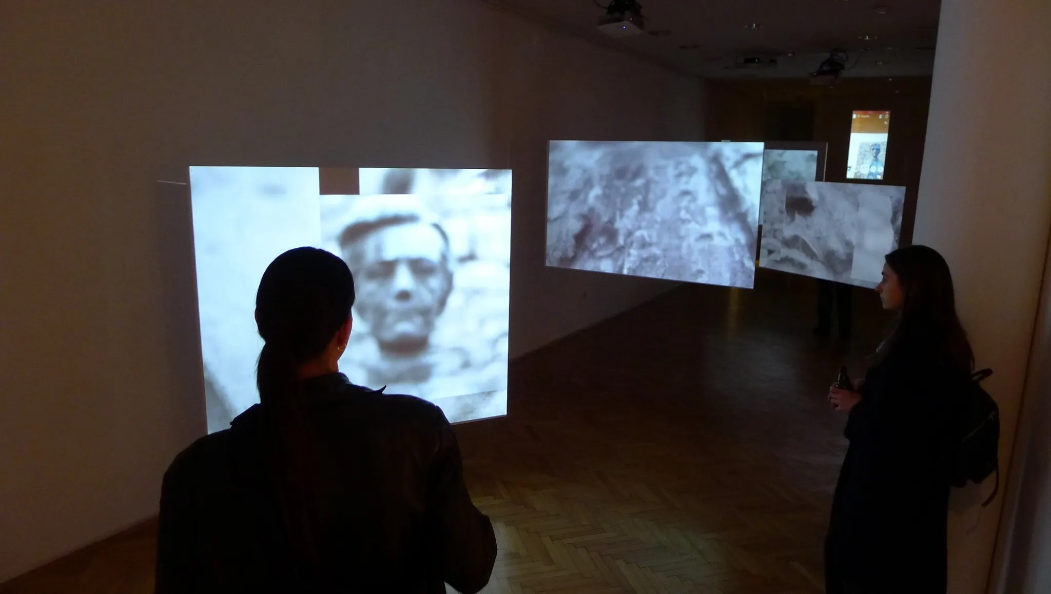 Installation view: visitors looking at multiple projected screens showing grainy close-up faces and stone textures.