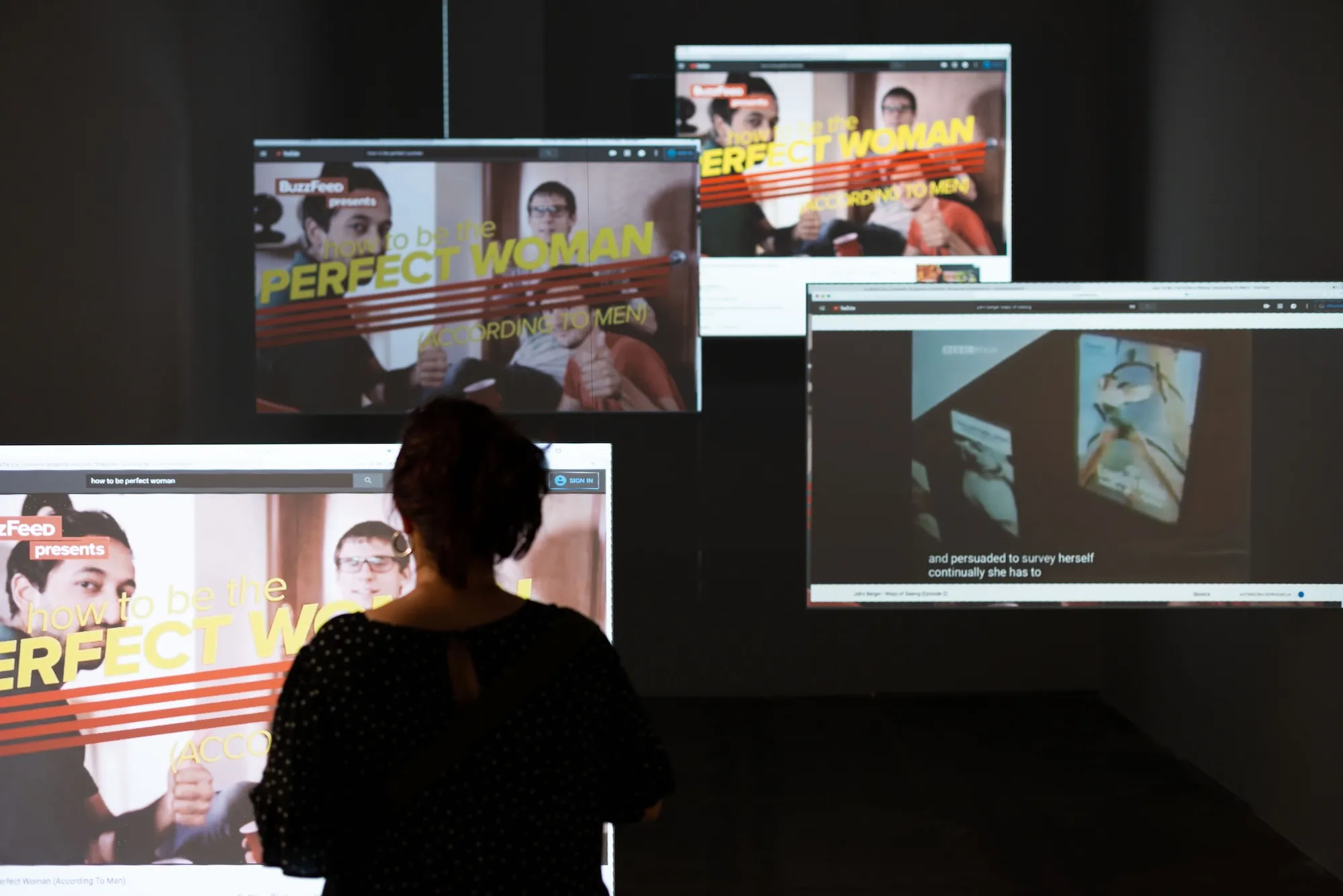 Installation view: a visitor's silhouette in front of multiple screens showing a BuzzFeed video titled 'How to Be the Perfect Woman (According to Men)'.