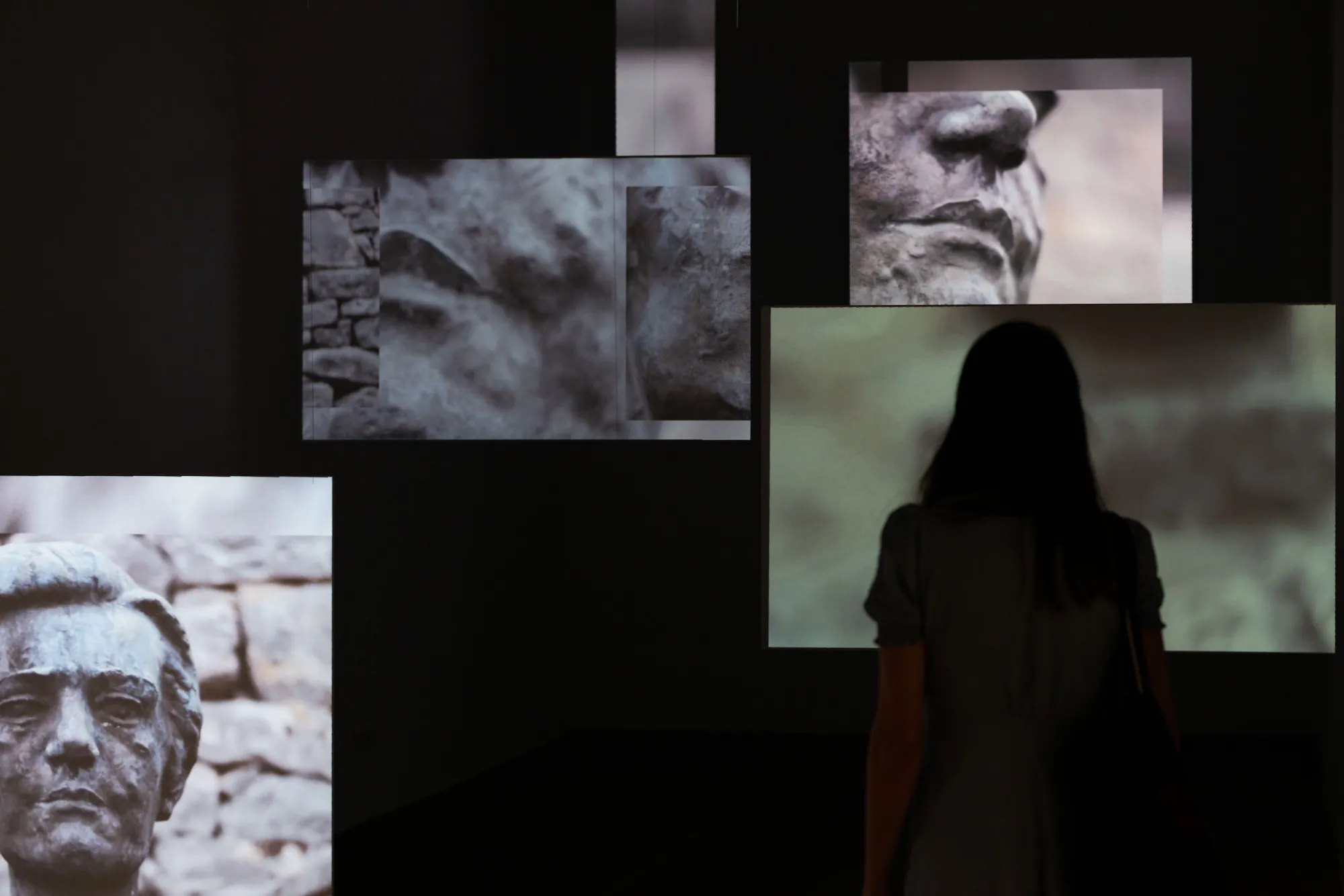 Installation view: multichannel screens showing close-ups of a weathered stone bust, with a visitor looking at a green-toned screen.
