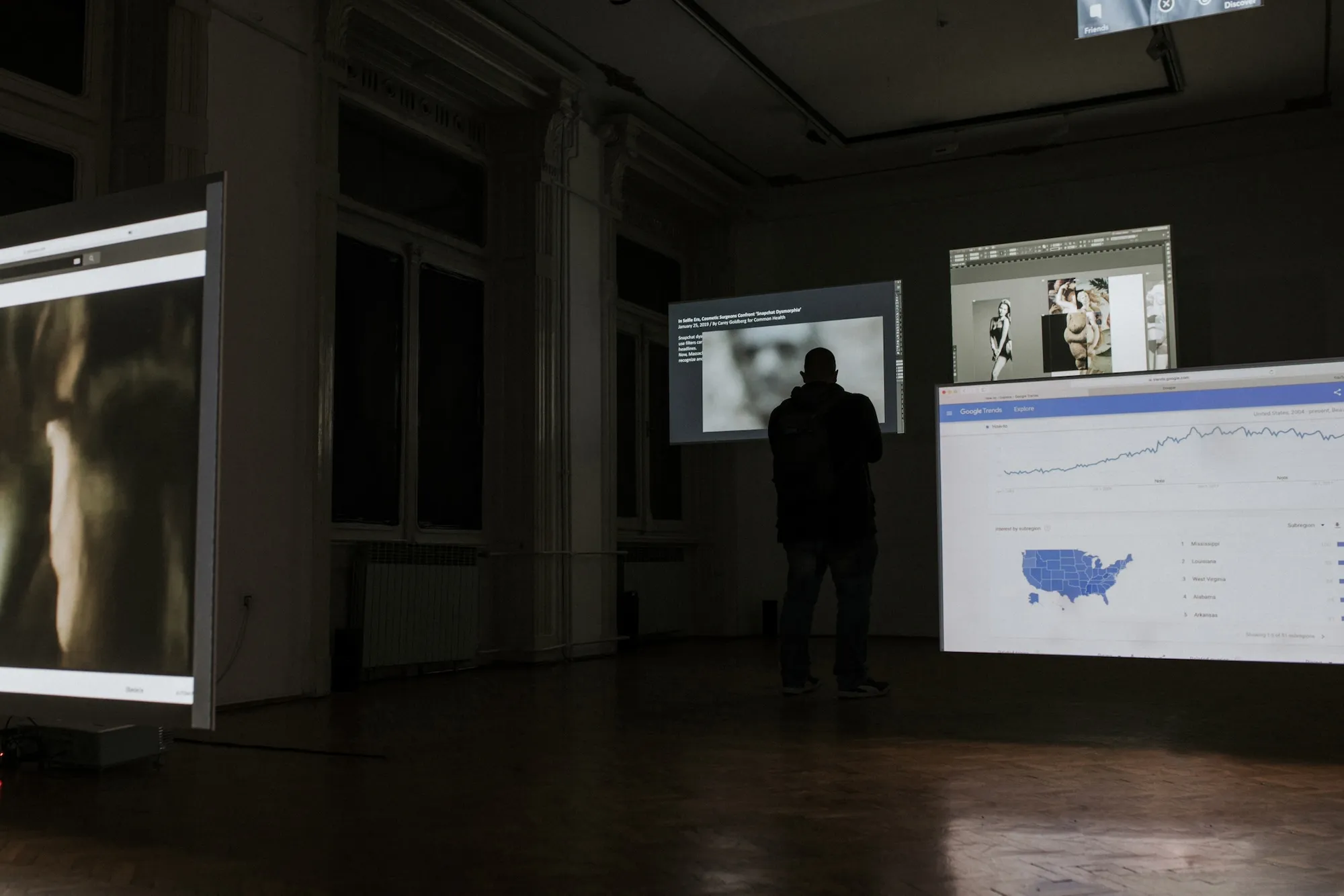 Installation view: a screen showing the article 'In Selfie Era, Cosmetic Surgeons Confront Snapchat Dysmorphia', beside a photo collage of classical nude figures, with a visitor standing in front.