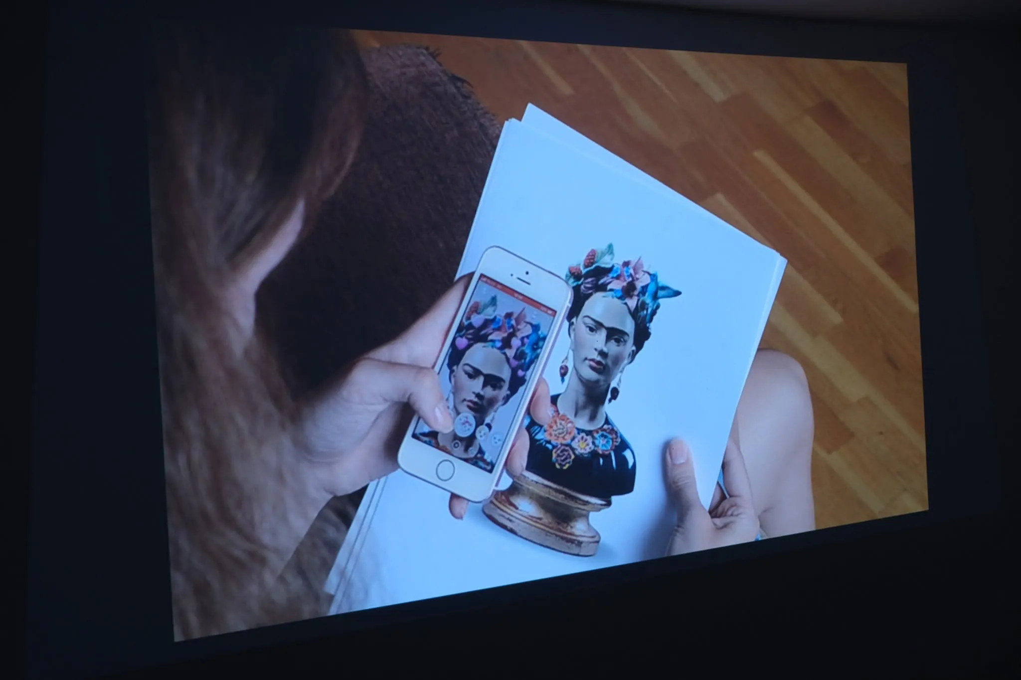 A projected video still of a hand holding a phone showing a Snapchat-filtered selfie of a Frida Kahlo bust, held up beside a printed photo of the same bust.
