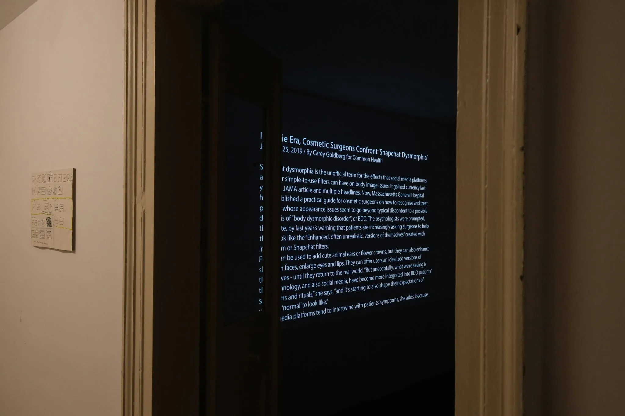 A dark corridor with a doorway showing a projected screen of the "Snapchat Dysmorphia" article text, with a wall didactic panel visible beside it.