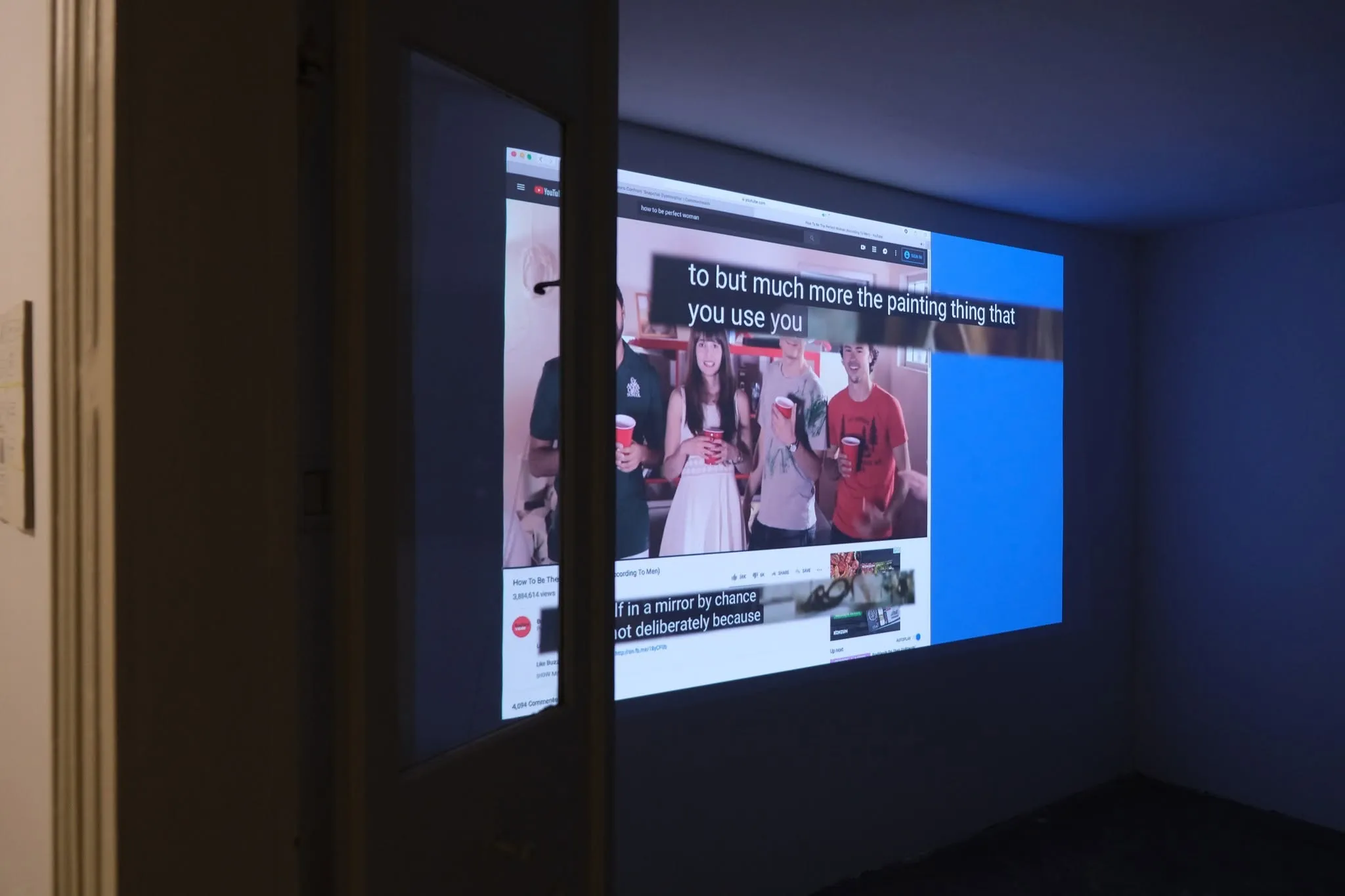 A dark room with a projected YouTube video, "How To Be The Perfect Woman (According To Men)", showing captions over a group of people holding red cups.