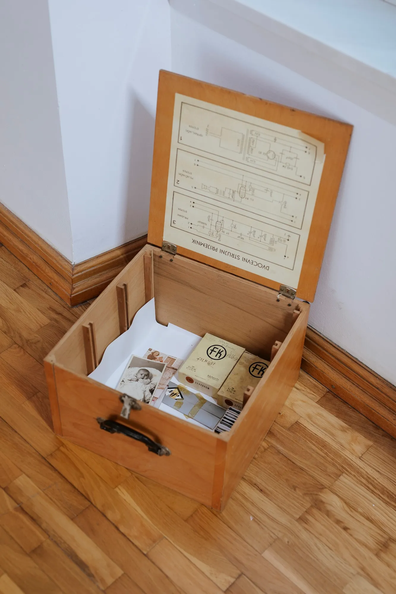 An open wooden box containing old photographs, labeled film boxes, and a printed diagram on the underside of its lid.