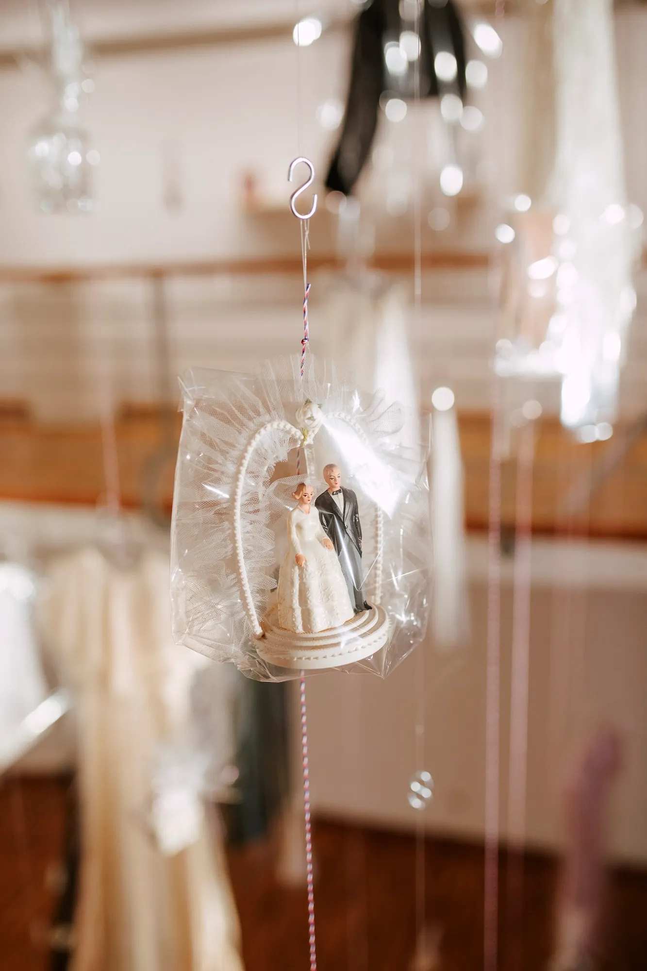 A miniature wedding cake topper of a bride and groom, wrapped in clear film and suspended by a string.