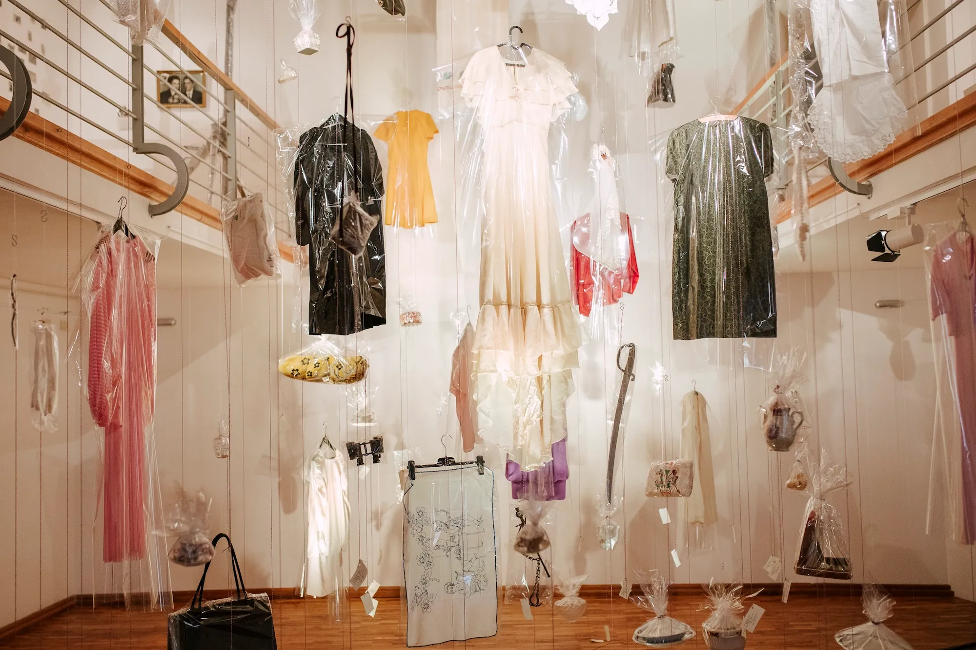 Wide installation view of dresses and everyday objects wrapped in clear Melinex museum archival film, suspended at varying heights across the gallery.