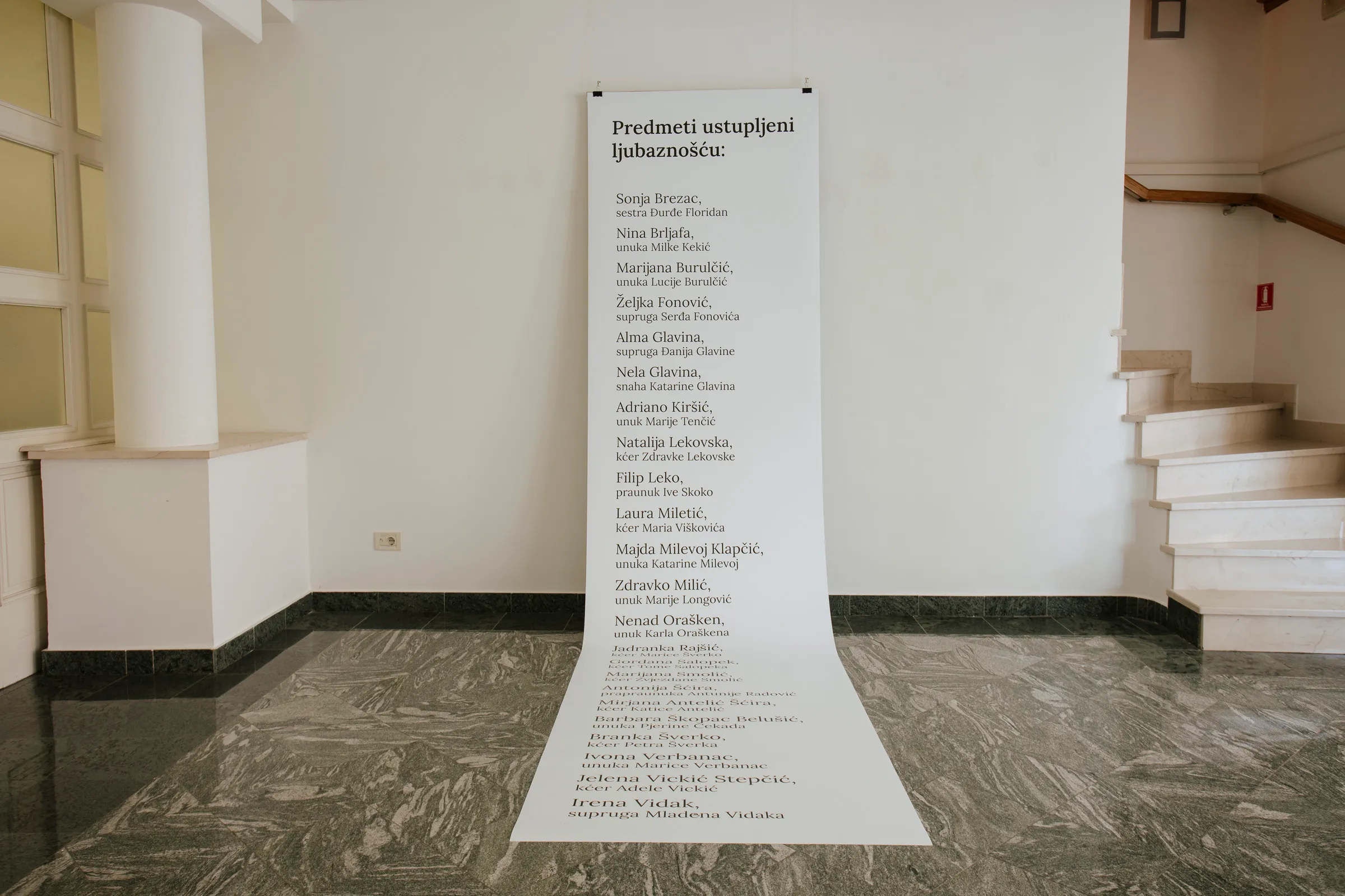 A long paper scroll listing donor names hanging from ceiling to floor in the gallery stairwell, with a staircase visible alongside.