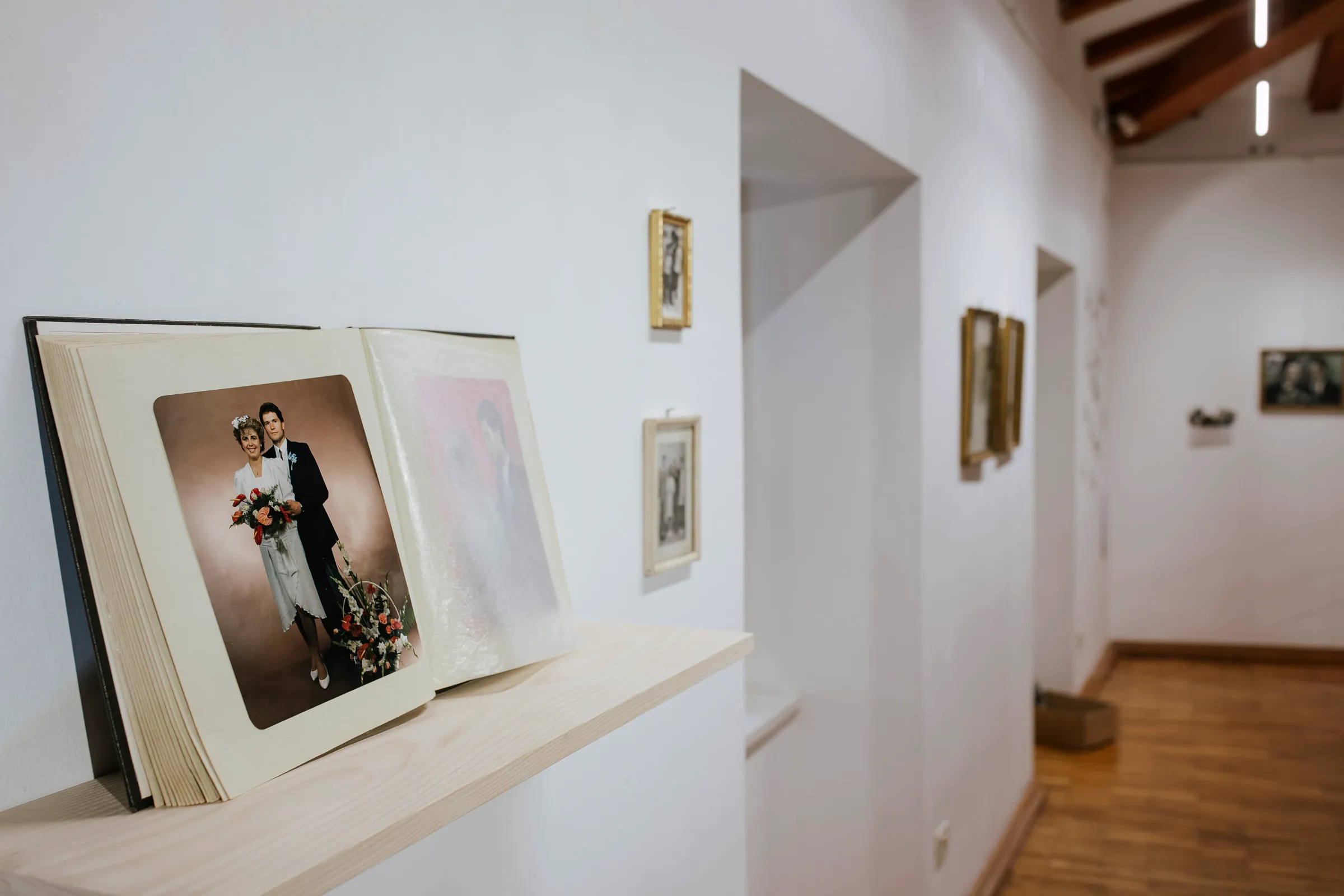 An open photo album showing a wedding portrait resting on a wooden shelf, with small framed photographs on the wall and a gallery hallway beyond.