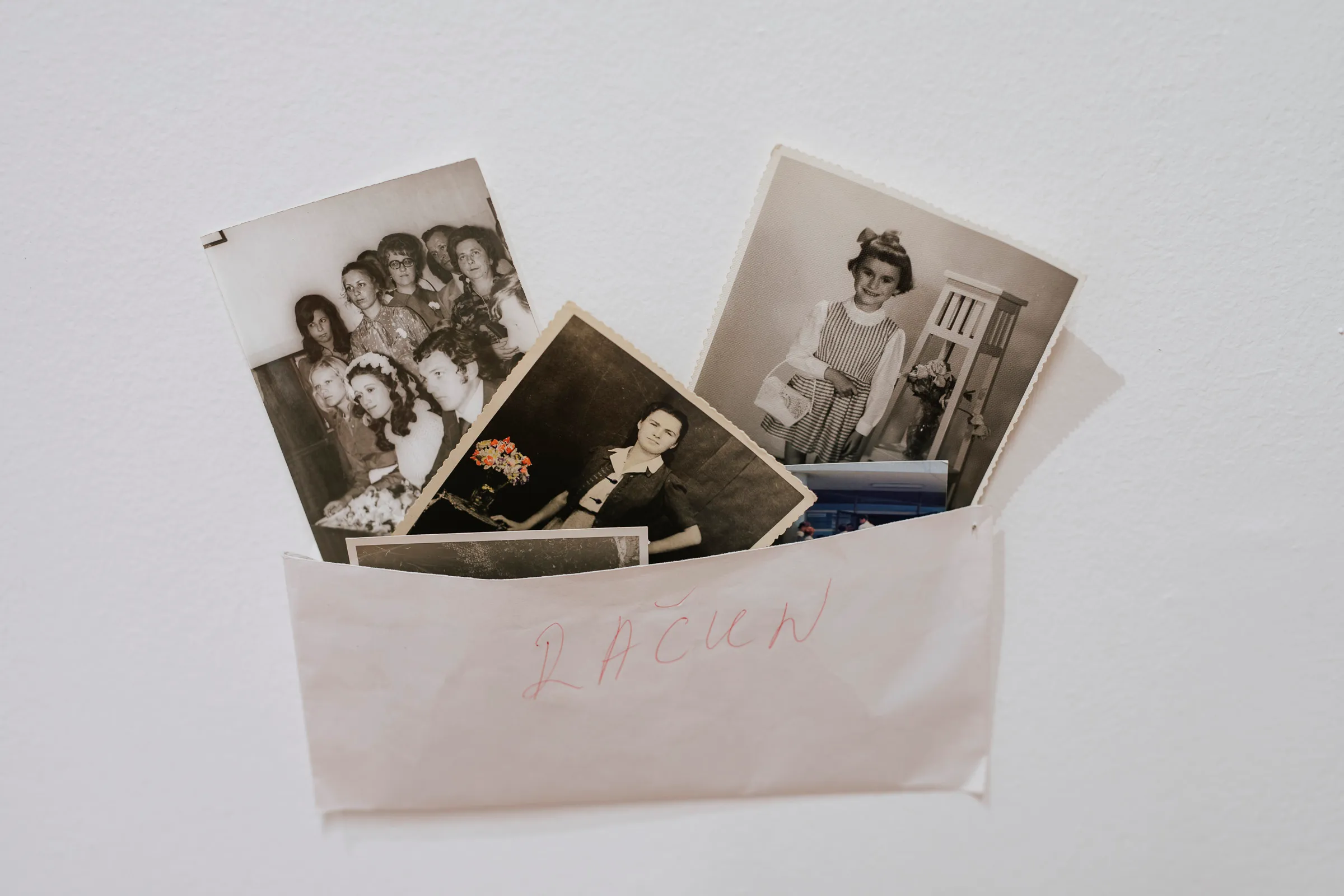 A paper envelope taped to the gallery wall holding several family photographs, including group portraits and a child's portrait, with a handwritten label in red.
