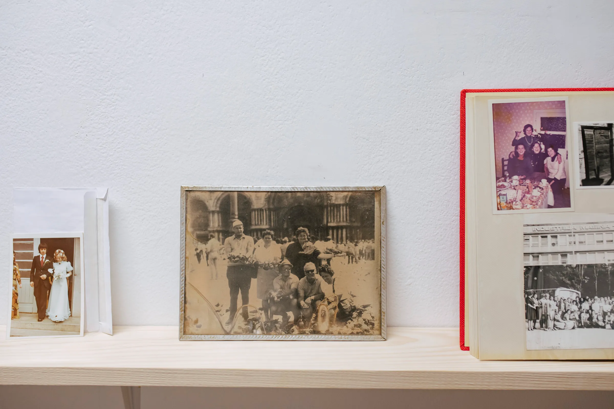 A shelf display of a wedding photo tucked in an envelope, a framed sepia group photograph, and an open red photo album with pasted family snapshots.