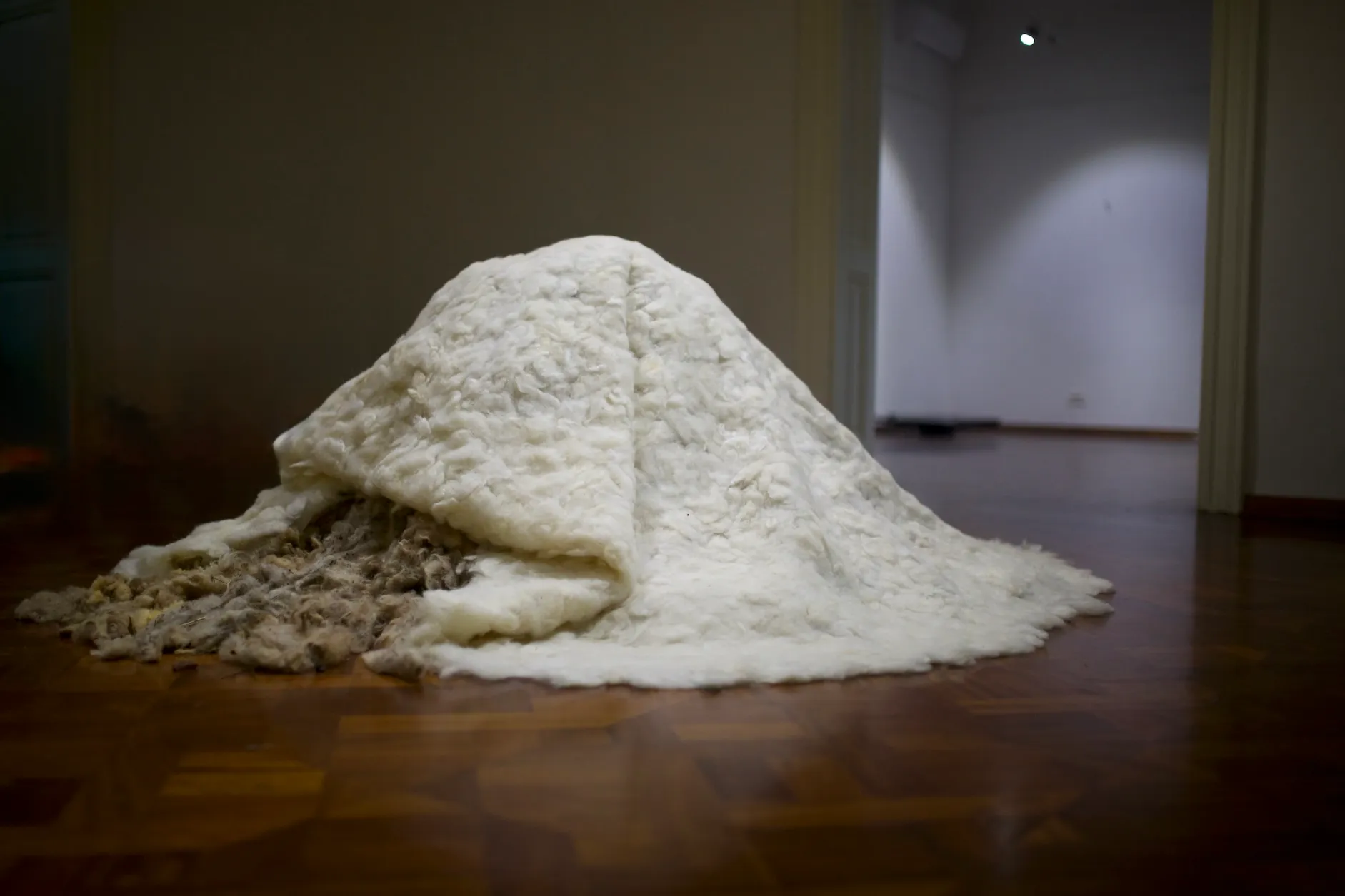 Installation view of a large mound of white felted wool spilling through a doorway into a room with a chair and a patterned rug.