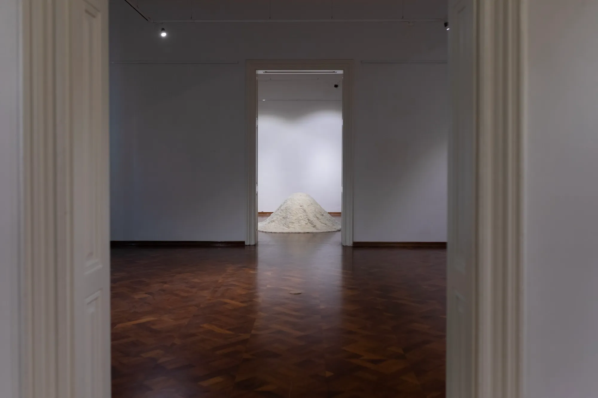 A framed doorway view through an empty gallery room to the mound of felted wool visible in a second room beyond.