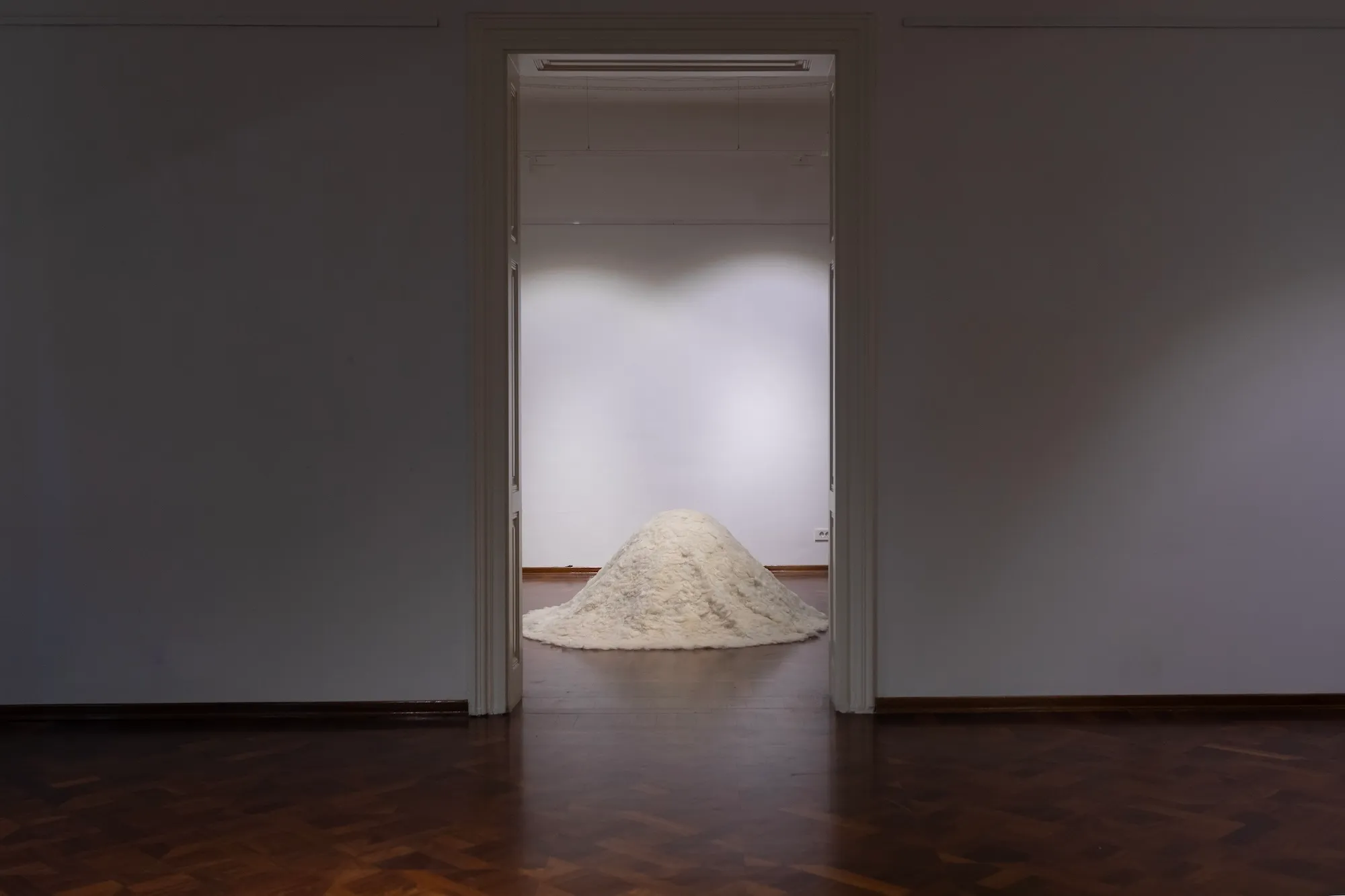 Distant symmetrical view through a doorway of the felted wool mound glowing under gallery light in the room beyond.